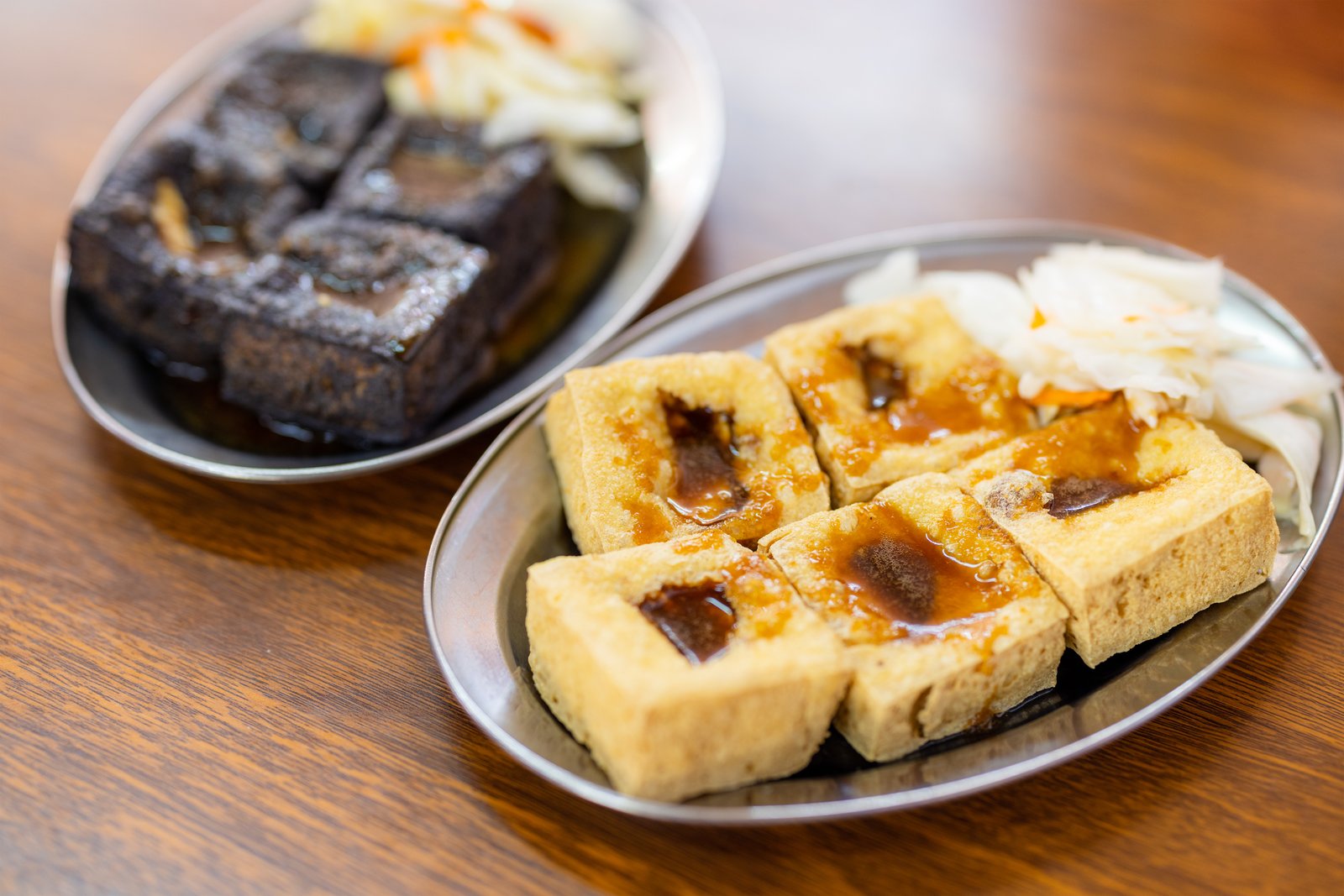 Two plates of stinky tofu, one deep black and one fried golden, served with sauce and pickles